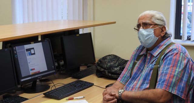An older gentleman wearing a face mask sits intently listening to his tutor at a computer desk at Moira adult learning centre.