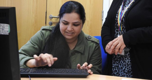 A learner in a green shirt is typing on a computer whilst her tutor watches and instructs.