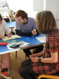 A maths learner is looking over as another learner on his table explains her working-out on a question. Learners are observed by tutor wearing red plaid dress.