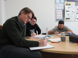 An image of three adults seated around a table in a classroom, writing in notebooks and taking part in a group learning activity.