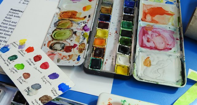 Watercolour paints are displayed out on a table with a learner's hand-made colour reference card visible.