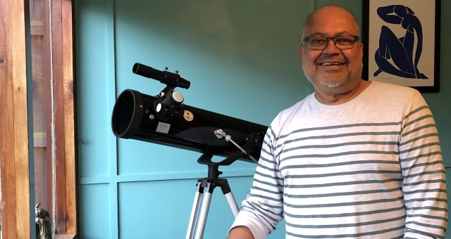 Photograph shows one of our astronomy learners at home with his telescope. He is wearing a white stripy jumper and is standing by an open window against a teal blue wall.