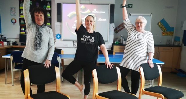 Two learners in white and grey tops are standing either side of their tutor. All three are demonstrating a yoga pose using chairs to aid balance.