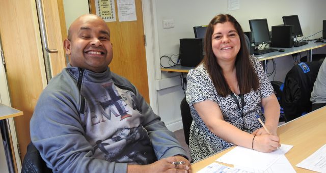 Learner is sitting at a desk with his learning support assistant and posing for the camera.