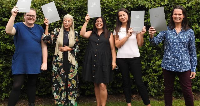 Image is taken outdoors at Moira Adult Learning Centre in front of the lush green bushes that surround the venue. In the image five learners hold up grey certificate folders in celebration of completing their course.