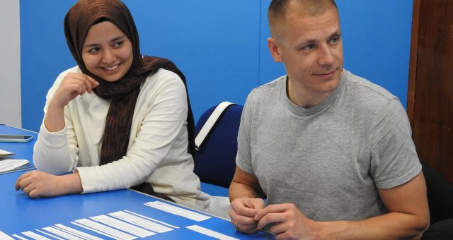 Two learners sit at a blue desk with a language game in front of them. They are looking off to the side of the image listening to their tutor.