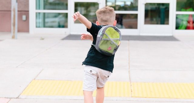 Young boy is wearing a green backpack and running excitedly towards the entrance to a school on his first day.