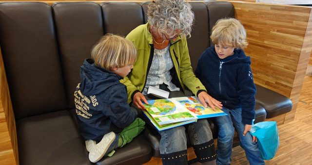 Image shows an older lady with white hair sitting between two young children reading a children's book.