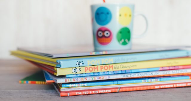Image shows a pile of colourful children's books in the foreground, a soft focus children's mug can be seen in the background.