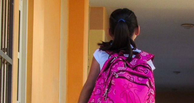 Young girl is wearing a bright pink backpack and walking towards the entrance of her school on her first day.