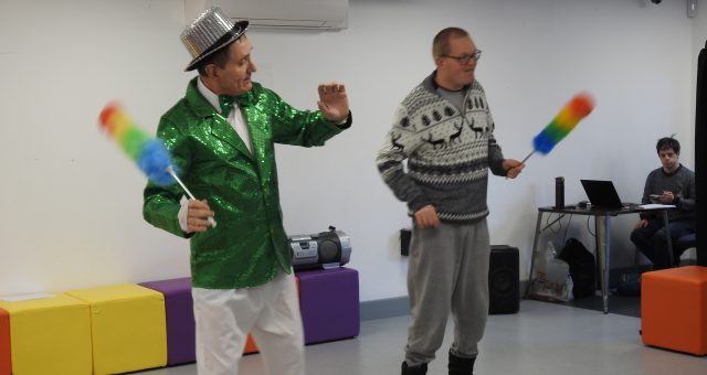 Two learners perform a dance to 'Happiness' by Ken Dodd, holding feather dusters and wearing costumes.
