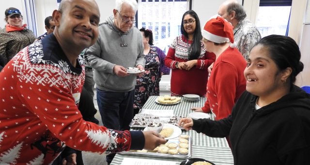Image shows a table laid with homemade mince pies and other treats, the table is surrounded by learners, staff, family and friends all wearing colourful Christmas jumpers. In the foreground two learners are posing for the camera smiling. One of the learners is pretending to hand the other a mince pie and a chocolate Krispy cake on a plate.