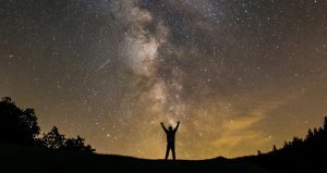 An image of a Silhouette of a person standing on a hill with arms raised beneath a star-filled night sky and the Milky Way stretching overhead.