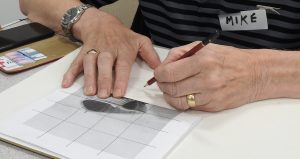 An image of a person seated at a table using a pencil and ruler to work on a drawing or shading exercise, with a smartphone and learning materials nearby.