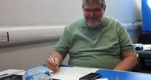 An image of a person seated at a table writing or sketching on paper during an art or learning activity, with a smartphone and art materials on the table.