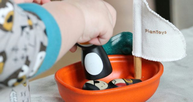 An image of Close-up of a young child’s hand placing a wooden play figure into an orange toy boat during a play-based learning activity.