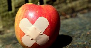 Red apple with two adhesive bandages crossed over it, placed outdoors on a wooden surface.