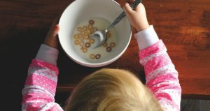 Overhead view of a young child eating cereal from a bowl at a wooden table while holding a spoon.