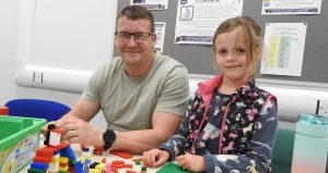 An adult and a child sit at a table taking part in a learning activity with colourful building blocks in a classroom setting.