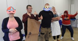 Group of adults taking part in a drama activity, wearing handmade costume props including masks and cardboard outfits while posing in a classroom or studio setting.