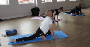An image of Adults taking part in a yoga class on exercise mats, performing a stretch in a bright studio with wooden flooring and large windows.