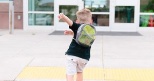 A young child wearing a backpack walks across a pavement outside a school entrance, with arms raised and school doors visible in the background.