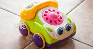 Colourful toy telephone with a smiling face, pink rotary-style dial and wheels, sitting on a tiled floor.