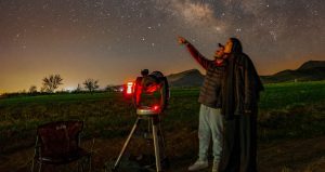 An image of a Person standing beside a telescope at night, pointing towards a star-filled sky while observing the night sky in an open outdoor landscape.