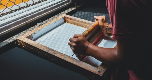 An image of a Person using a screen-printing frame and squeegee to apply ink across a mesh screen in a printmaking workshop.