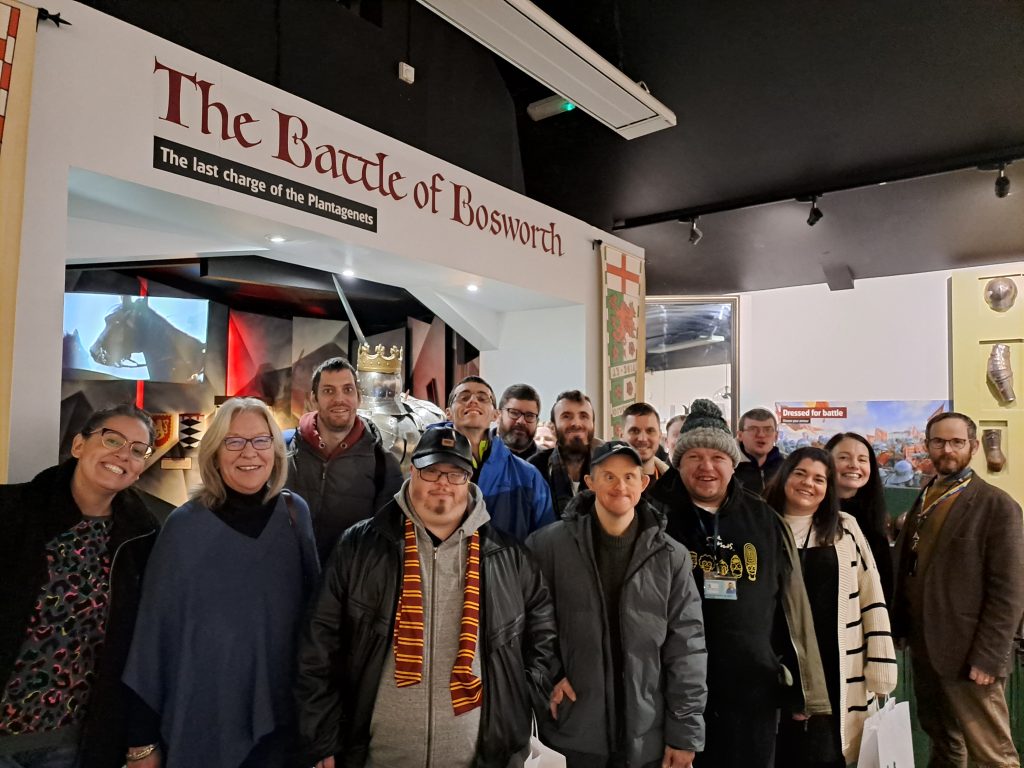 Image shows a large group of learners and staff smiling at the camera Infront of a display commemorating the Battle of Bosworth.