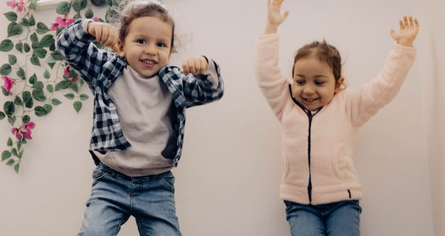 An Image of two young children jump in the air indoors above a small toy train on a wooden floor, with a window and decorative flowers in the background.