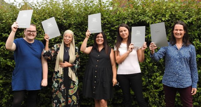Five adults standing outdoors in front of a hedge, each holding up a certificate folder during a celebration of learning or achievement.