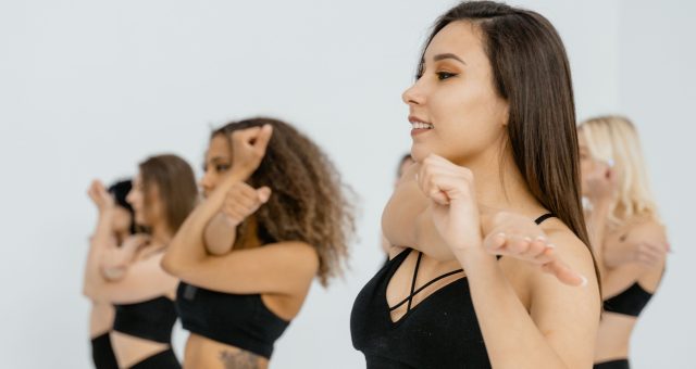 Group fitness class performing coordinated dance-based exercise movements in a bright studio, with participants standing in rows wearing activewear.