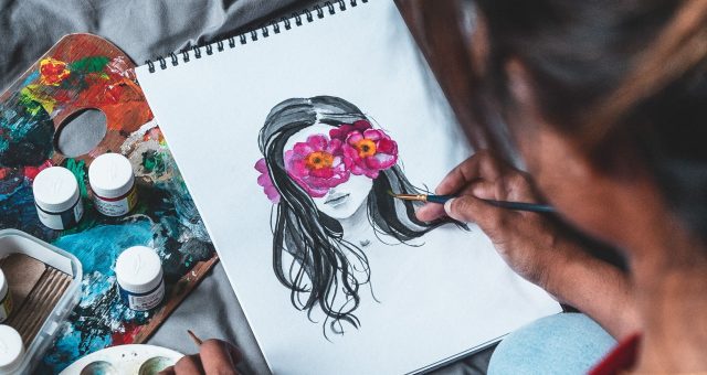 Person painting a portrait illustration in a sketchbook, using a fine paintbrush while surrounded by paints, palettes and art materials.