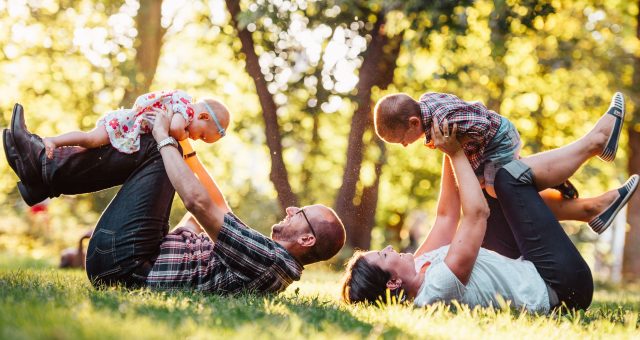 An image of two adults and two young children play together on grass in a sunlit park, with the adults lifting the children above them while lying on the ground.