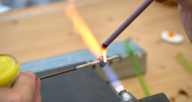 An image of a close-up of hands using a small torch flame to heat and shape molten glass on metal rods during a glassmaking activity.
