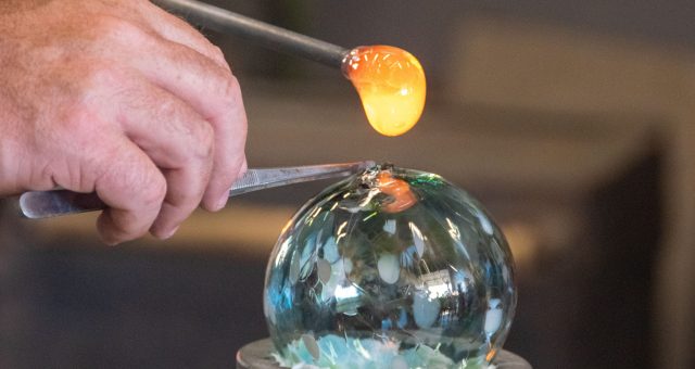 An Image of a close-up of a glassmaking process, with a glowing piece of molten glass being applied to a decorative glass sphere using metal tools.