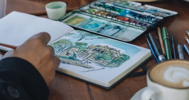 An image of an open sketchbook displaying a pen-and-watercolour illustration of a house and garden, with paint palette, drawing pens and a cup of coffee on a wooden table.