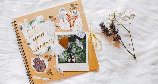 An image of Decorated scrapbook with photographs, stickers, feathers and dried flowers arranged on a white fabric background.