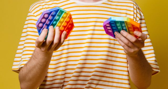 Person holding two colourful rainbow pop-it sensory toys against a bright yellow background.