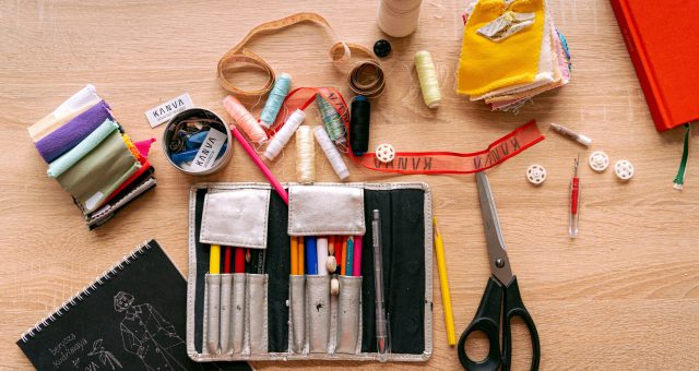 An image of sewing and textile craft materials arranged on a wooden table, including fabric samples, thread reels, measuring tape, scissors, buttons, pencils and a sketchbook.