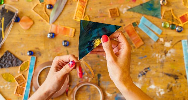 An image of a close-up of hands working with coloured glass pieces on a craft table, wrapping copper foil around a triangular piece of blue glass during a stained-glass project.
