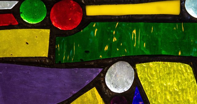 An image of a close-up of an abstract stained-glass panel featuring brightly coloured geometric shapes in green, yellow, red, blue, purple and white separated by dark lead lines.