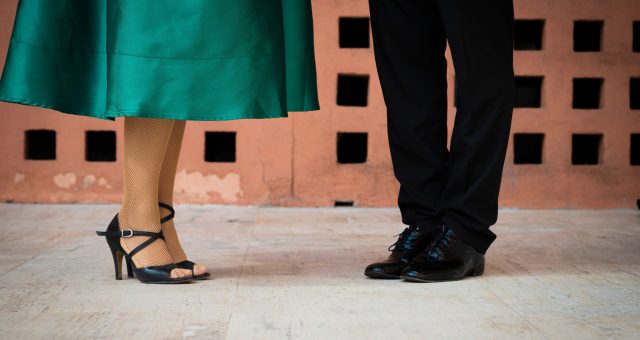 An image of Two people standing opposite each other in dance footwear, showing a dance position with feet visible against a patterned wall and light-coloured floor.
