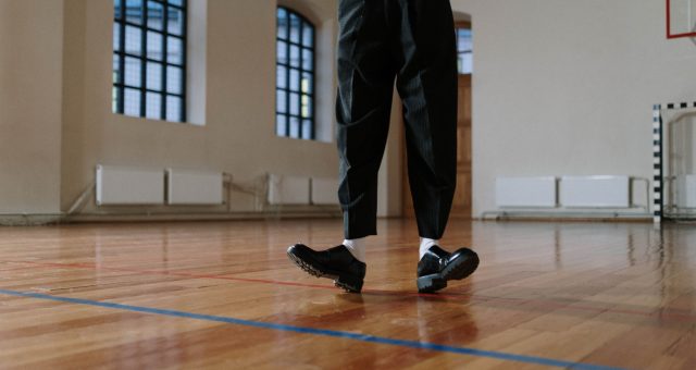 Person wearing tap shoes practising footwork on a polished wooden floor in a dance studio with large arched windows in the background.