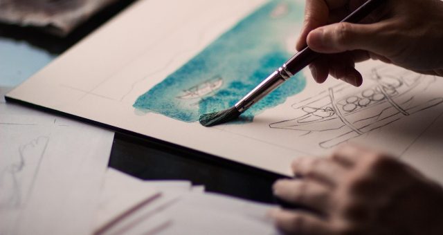 An image of a hand using a paintbrush to apply blue watercolour paint to a sketch on paper during a painting activity.
