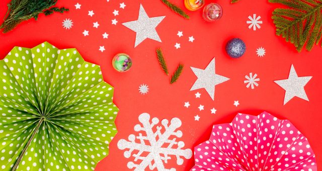 Festive craft display featuring folded paper decorations, silver stars and snowflakes, evergreen sprigs and colourful ornaments arranged on a bright red background.