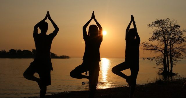 An image of three people practising yoga in tree pose beside a lake at sunset, silhouetted against the setting sun with hands pressed together above their heads.
