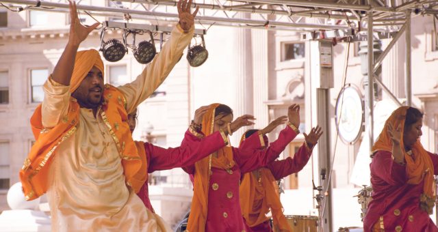 An image of a group of performers in colourful traditional costumes dancing on an outdoor stage, with raised arms and stage lighting equipment visible overhead.