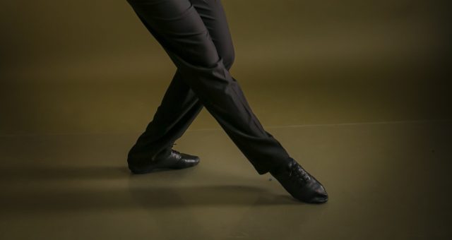 Performers legs in a black costume striking a dramatic dance pose against a plain studio background.